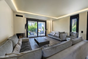 A modern living room with a sectional sofa, a coffee table, and a sliding glass door leading to an outdoor patio. - Grand Kourouta Pool Apartments (Ilida)