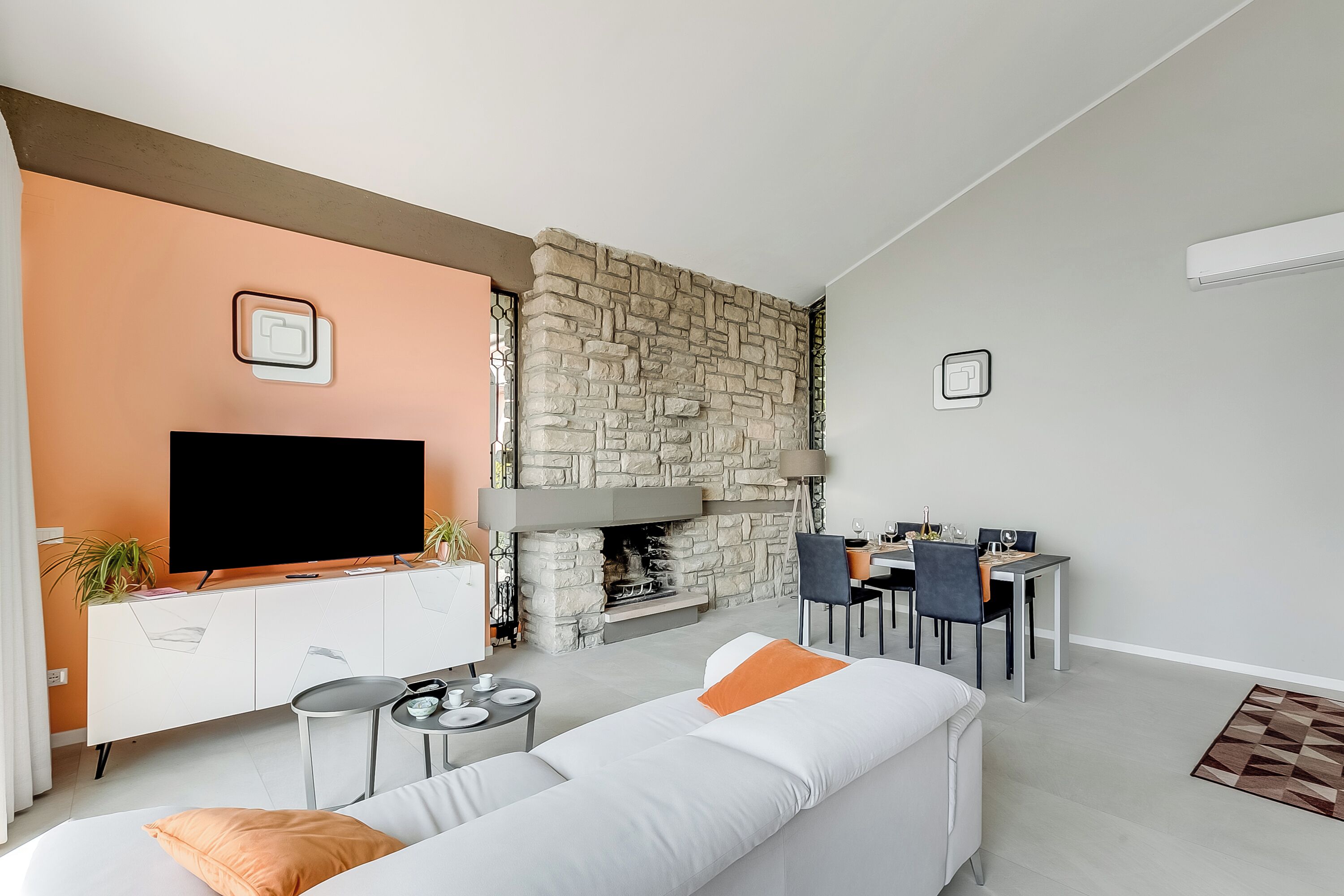 A modern living room with a stone fireplace, a flat-screen TV, a dining area, and a sofa with orange cushions.