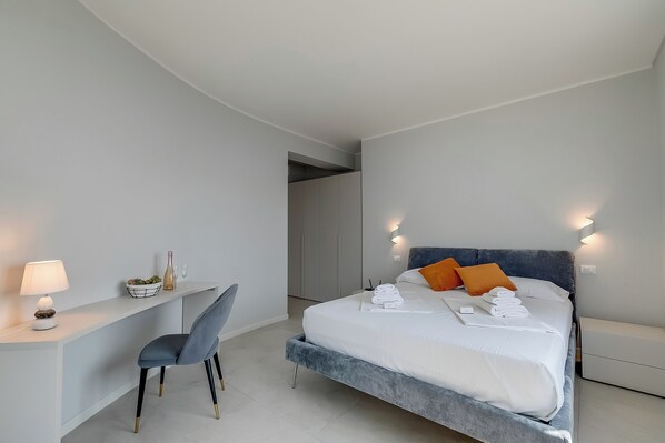 A modern hotel room with a bed, bedside table, chair, and a desk with a lamp.