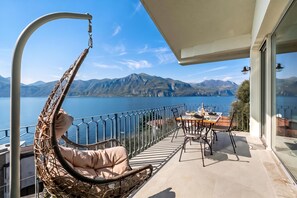 A balcony with a wicker chair, a table with chairs, and a view of a lake and mountains. - Villa Mercurio Apartments (Brenzone sul Garda)