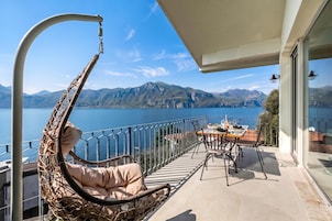 Un balcón con una silla de mimbre, una mesa y sillas, y vistas a un lago y montañas.