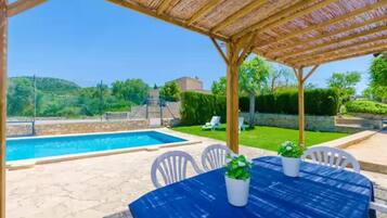 A covered outdoor area with a pool, white chairs, and a table with potted plants.