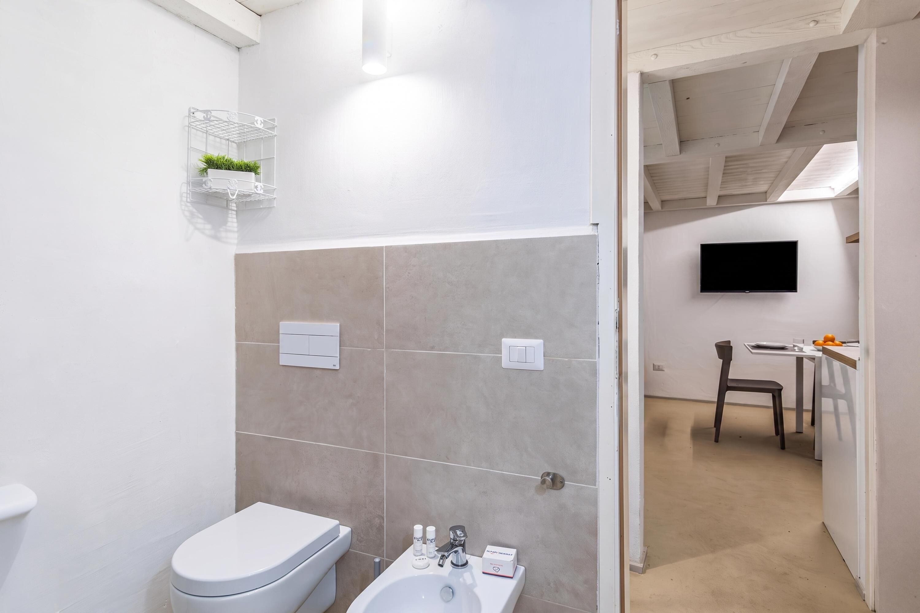 Egy modern fürdőszoba, amelyben WC, bidé és mosdókagyló található, egy kis polcon pedig egy növény, a szomszédos helyiségben pedig egy televízió.