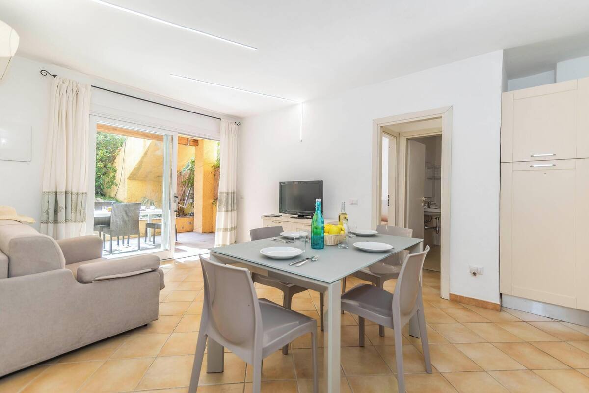 Una sala de estar moderna con comedor, un sofá, un televisor y una cocina visible a través de una puerta abierta.