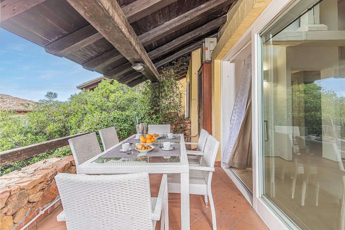An outdoor patio with a table set for two, surrounded by white wicker chairs, a stone wall, and a glass door leading to a balcony.