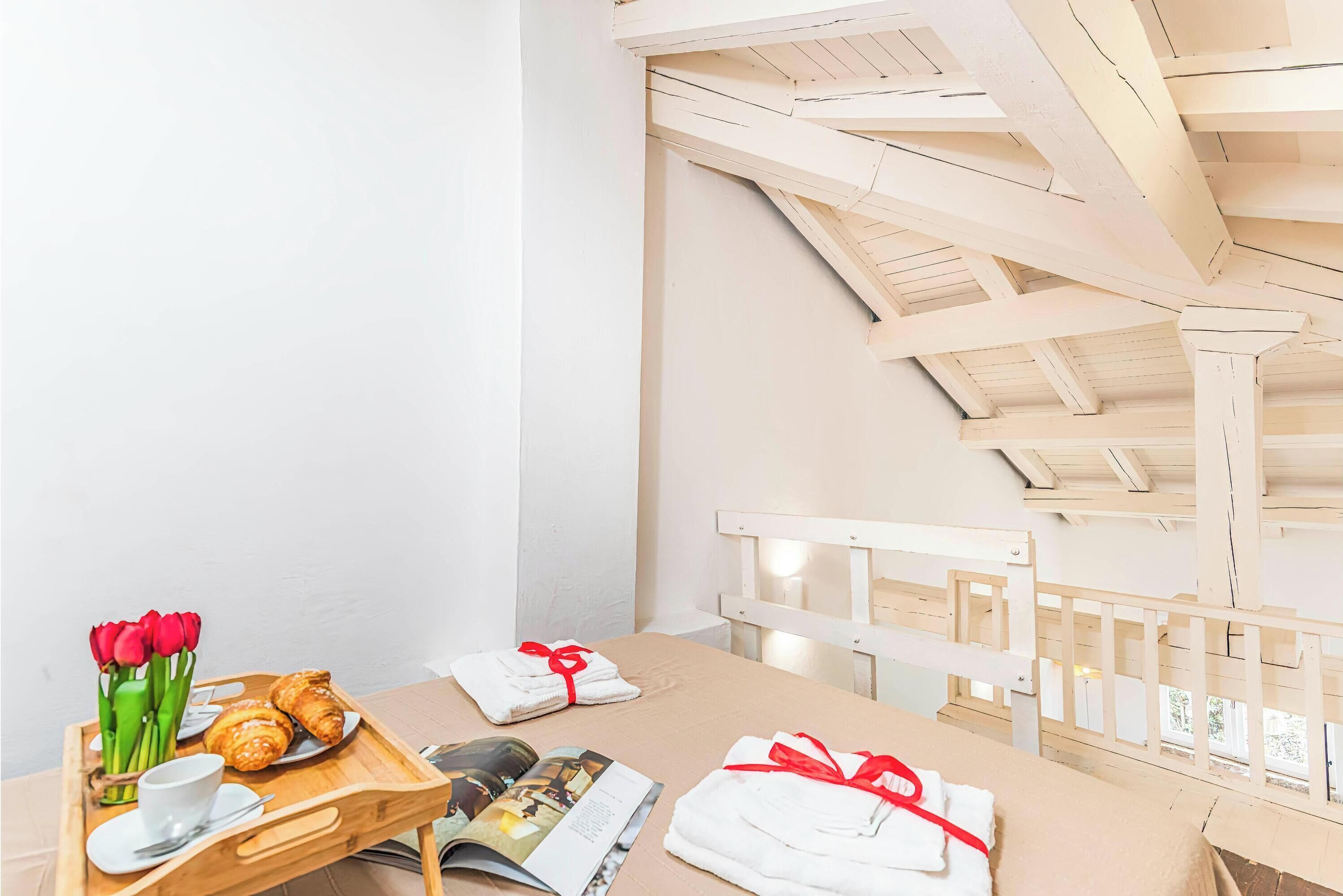 A room with a wooden ceiling, a bed with white bedding, a tray with croissants and a cup, and a magazine.