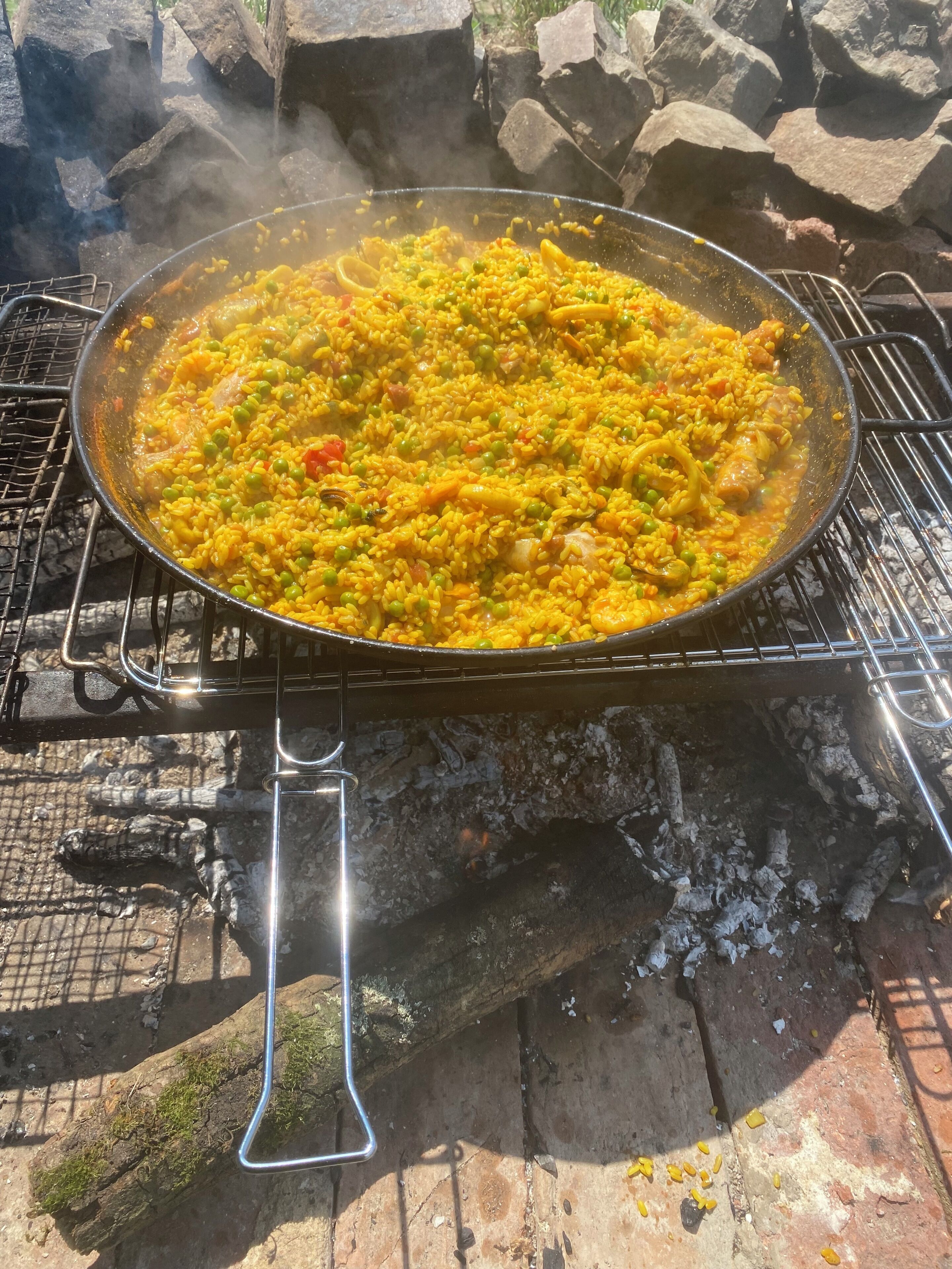 Een grote kookpot op een buitenbarbecue, met een metalen handvat, omringd door rotsen en een metalen gaas.