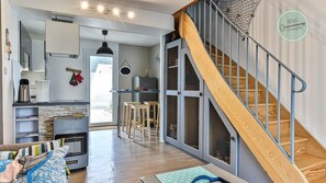 A modern kitchen with a built-in stove, a dining area with chairs, and a staircase with a wooden railing. - Maison Saint Surin 26 (Saint-Hilaire-de-Riez)