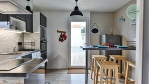 A modern kitchen with a built-in oven, microwave, and stainless steel countertops. There are wooden stools and a small table with a TV. - Maison Saint Surin 26 (Saint-Hilaire-de-Riez)