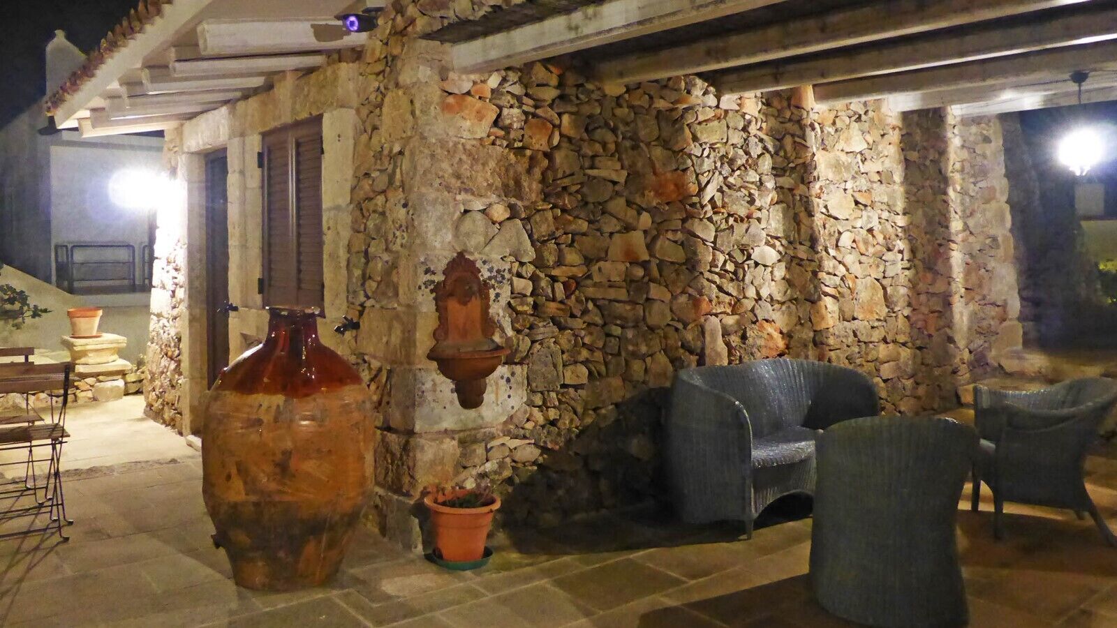 Edifício de pedra com fachada rústica, área de estar ao ar livre com cadeiras e um grande vaso de barro.