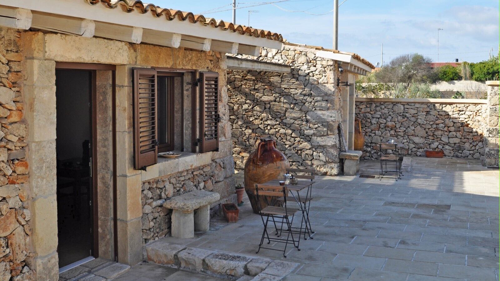 A stone building with a patio area featuring a large ceramic pot, outdoor seating, and a stone wall.