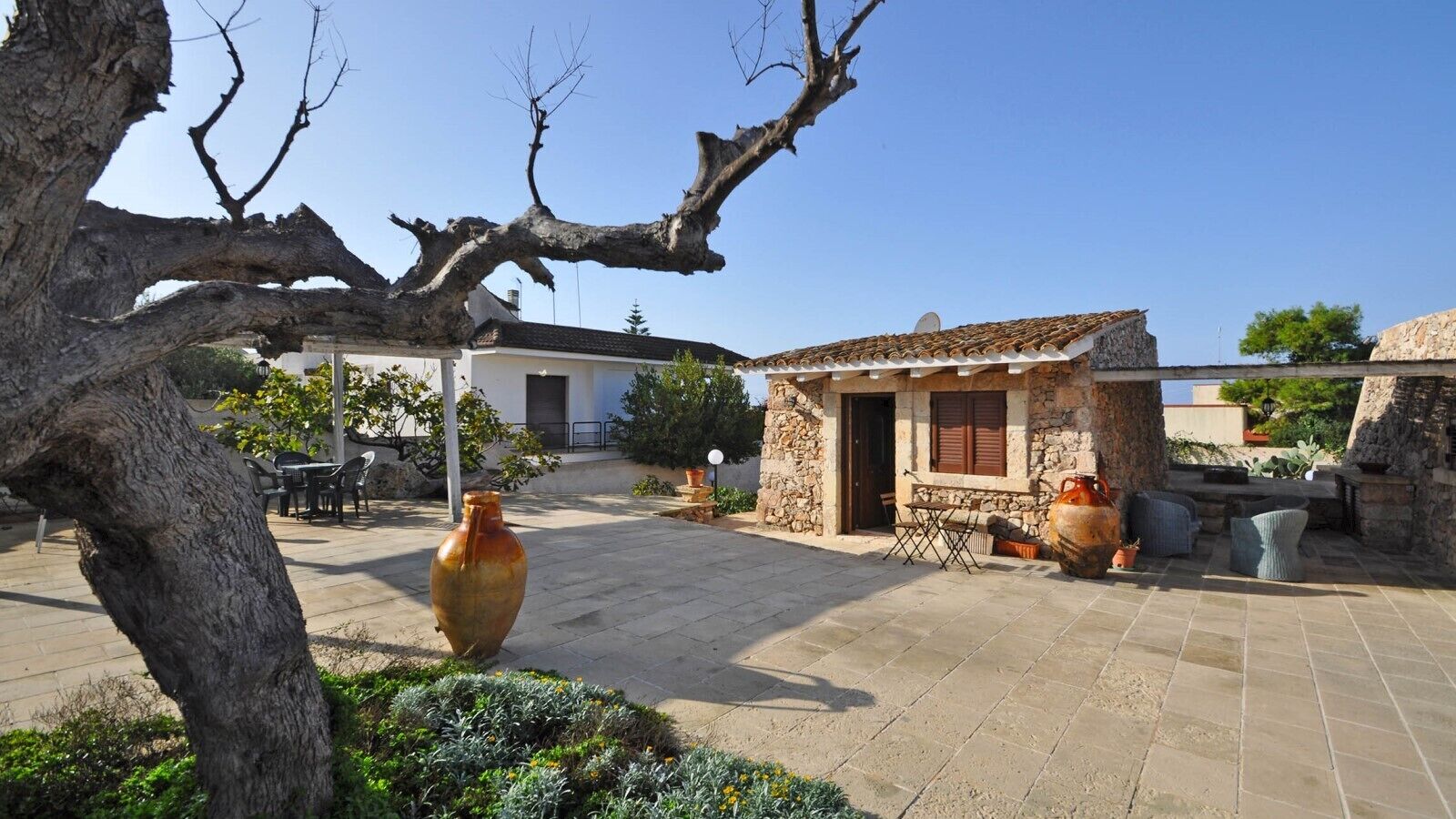 Un cortile con pavimento in pietra, un piccolo edificio in pietra con tetto in tegole, mobili da esterno e grandi vasi.