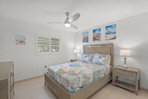 Une chambre à coucher comprenant un lit, deux tables de chevet, un ventilateur de plafond et des œuvres d’art accrochées aux murs.
