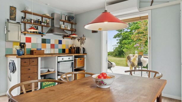 A kitchen with wooden cabinets, a dining table with chairs, a hanging red light fixture, and a view of a garden with a cow. - Holyday home in beatiful Vang (Hasle)
