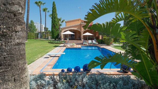 Ein Swimmingpool mit Liegestühlen und Sonnenschirmen, umgeben von Palmen und einem gepflegten Rasen.
