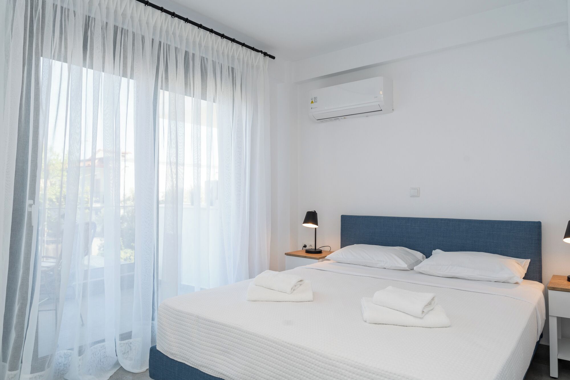 Une chambre d’hôtel avec un grand lit, du linge de lit blanc, une tête de lit bleue, un climatiseur, une lampe de chevet et des rideaux en voile.