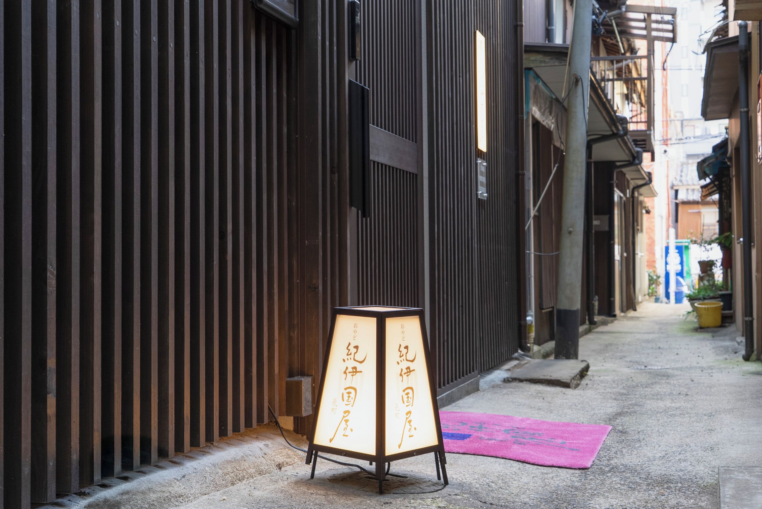 A narrow alley with a traditional Japanese lantern and a pink mat on the ground.