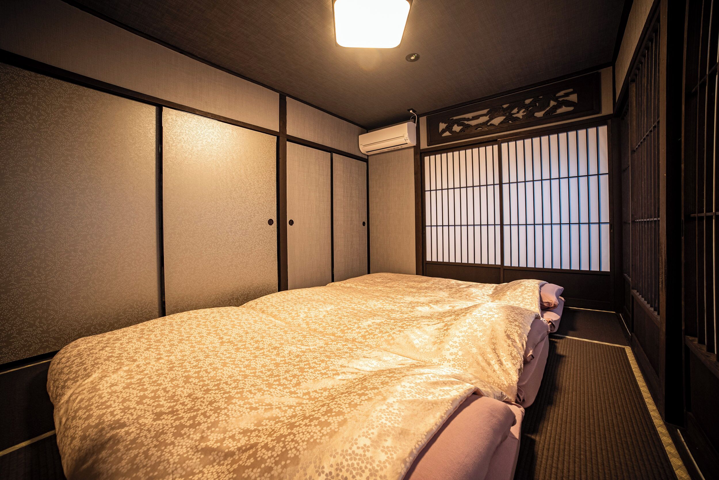 Ein traditioneller japanischer Raum mit Tatami-Boden, einem niedrigen Bett mit gemustertem Überwurf, Schiebetüren und einem Fenster mit Shoji-Bildschirm.