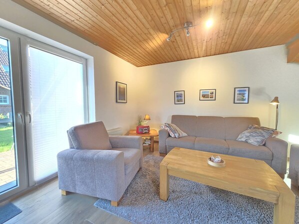 A modern living room with a wooden ceiling, a grey sofa, a wooden coffee table, and a large window with blinds.