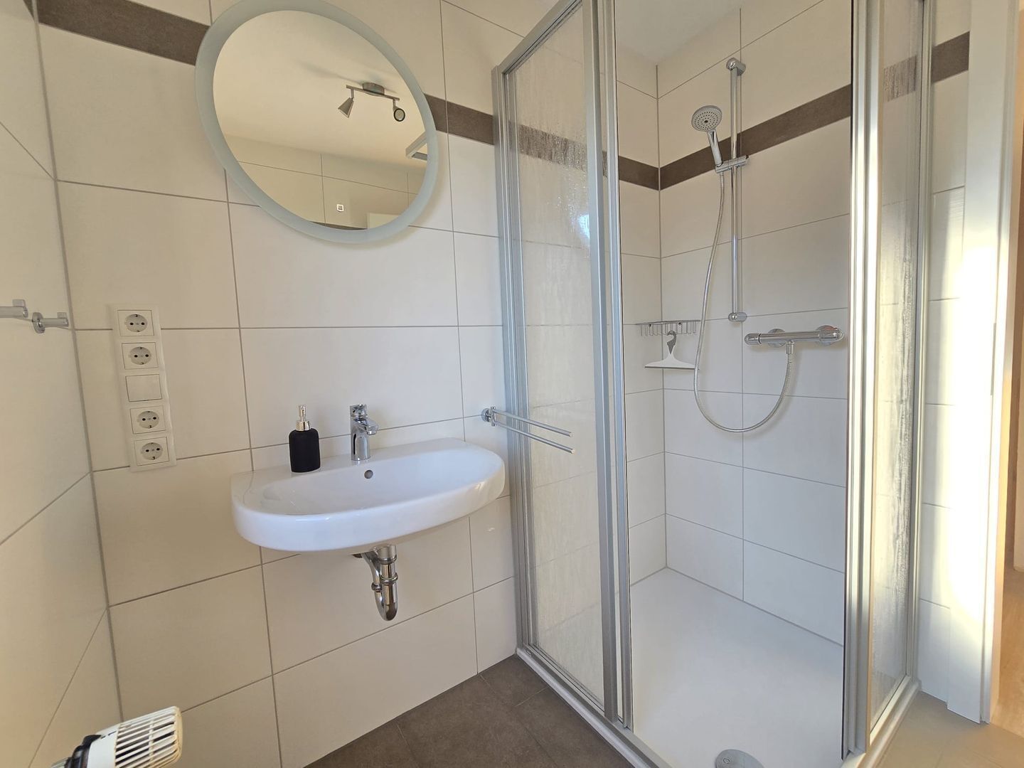 Une salle de bain avec une douche à parois de verre, un lavabo blanc avec un miroir au-dessus, et un sol carrelé.