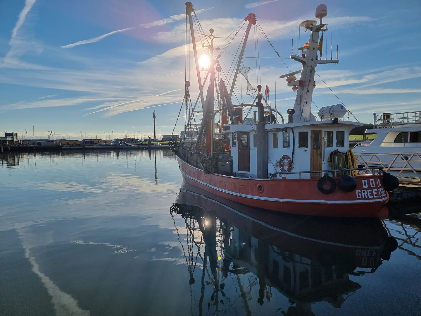 Een rode vissersboot afgemeerd in een jachthaven, met kalm water dat het schip en de lucht weerspiegelt.