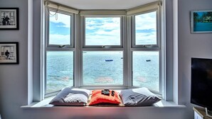 A bay window with a view of the sea, a bed with pillows, and a television on a stand.