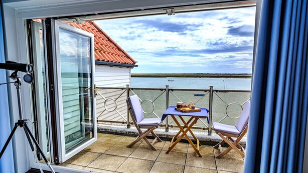 A balcony with a table and two chairs, overlooking a body of water and a distant shoreline.