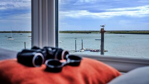 A view from a window overlooking a body of water with boats, a bird on a post, and a pair of goggles on a cushion.