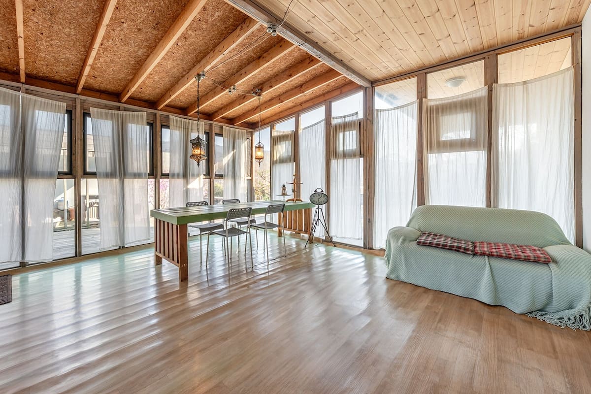 Ein geräumiges Zimmer mit Holzböden, einem Esstisch mit Stühlen, einem grünen Sofa und großen Fenstern mit leichten Vorhängen.