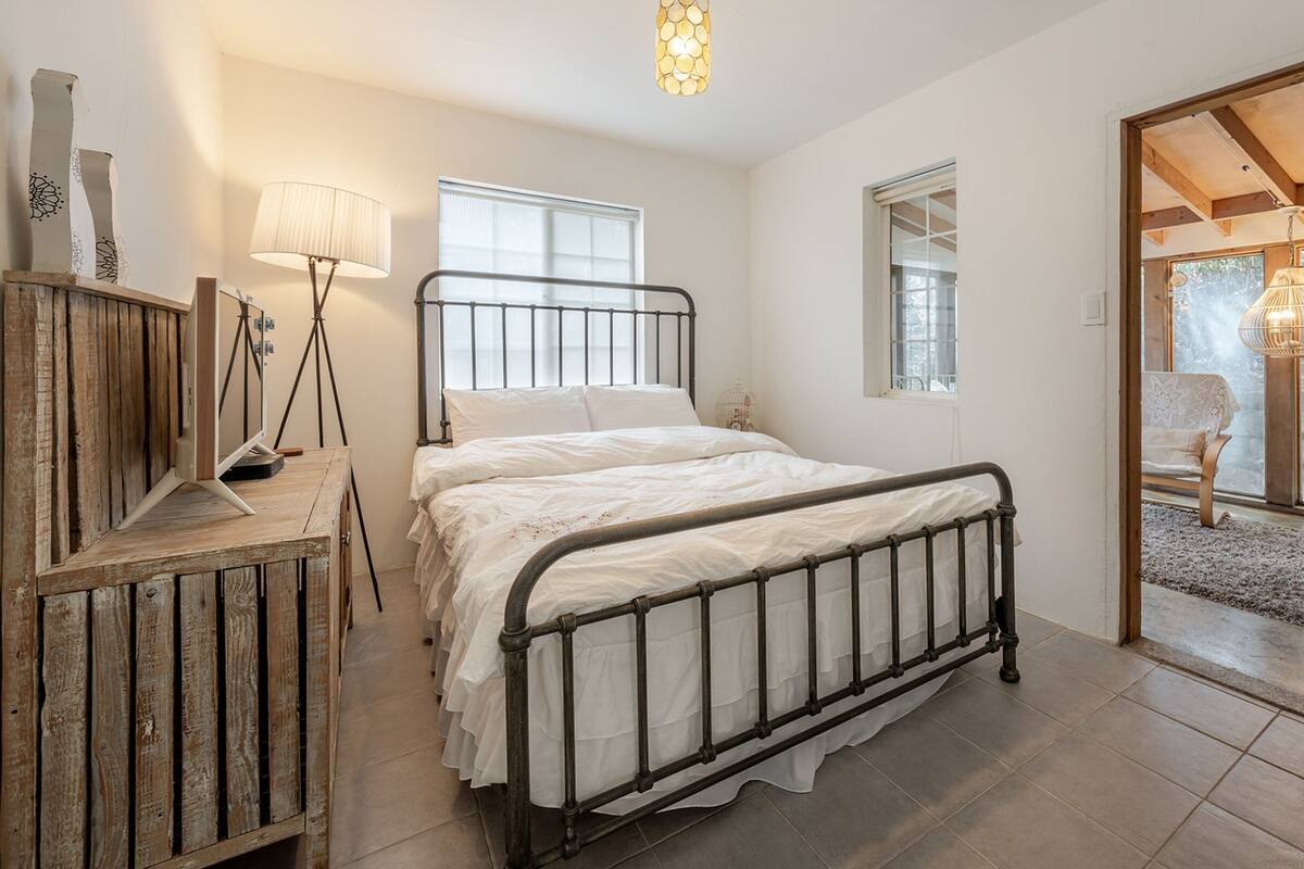 Ein Schlafzimmer mit einem Bett, einer Holzkommode, einer Lampe und Blick ins Freie.