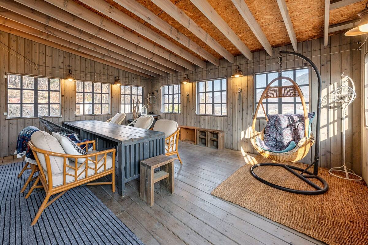 Ein Holzinterieur mit Esstisch, Stühlen, einem Hängekorbstuhl und großen Fenstern.