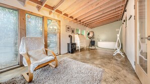 Een gezellige kamer met een houten plafond, een groot raam, een witte stoel, een witte dressoir, een badkuip en een witte vloerkleed.