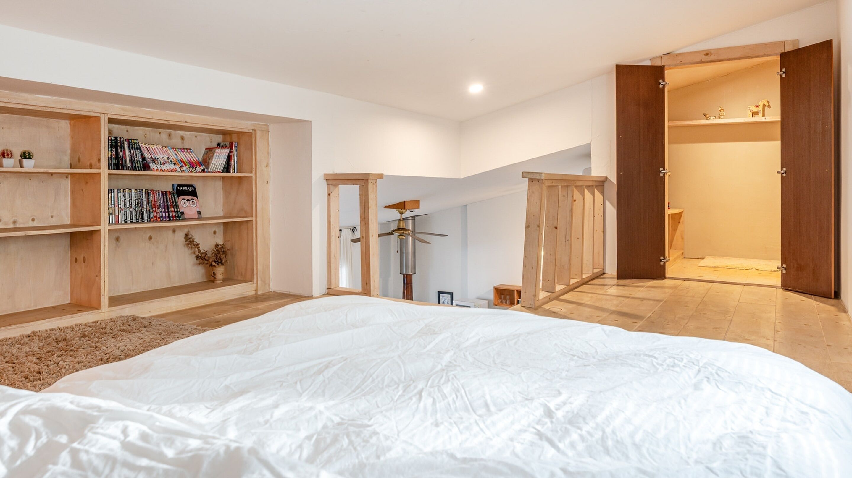 A bedroom with a large bed, wooden shelving with books and decorative items, a ceiling fan, and a wooden door leading to another room.