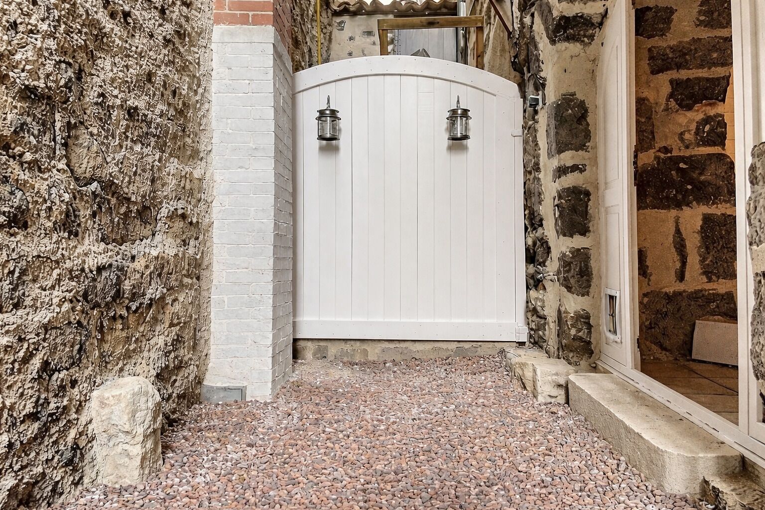 Une porte blanche, flanquée de deux lanternes, se dresse devant un mur de pierres et une clôture blanche, et mène à un bâtiment en pierre.