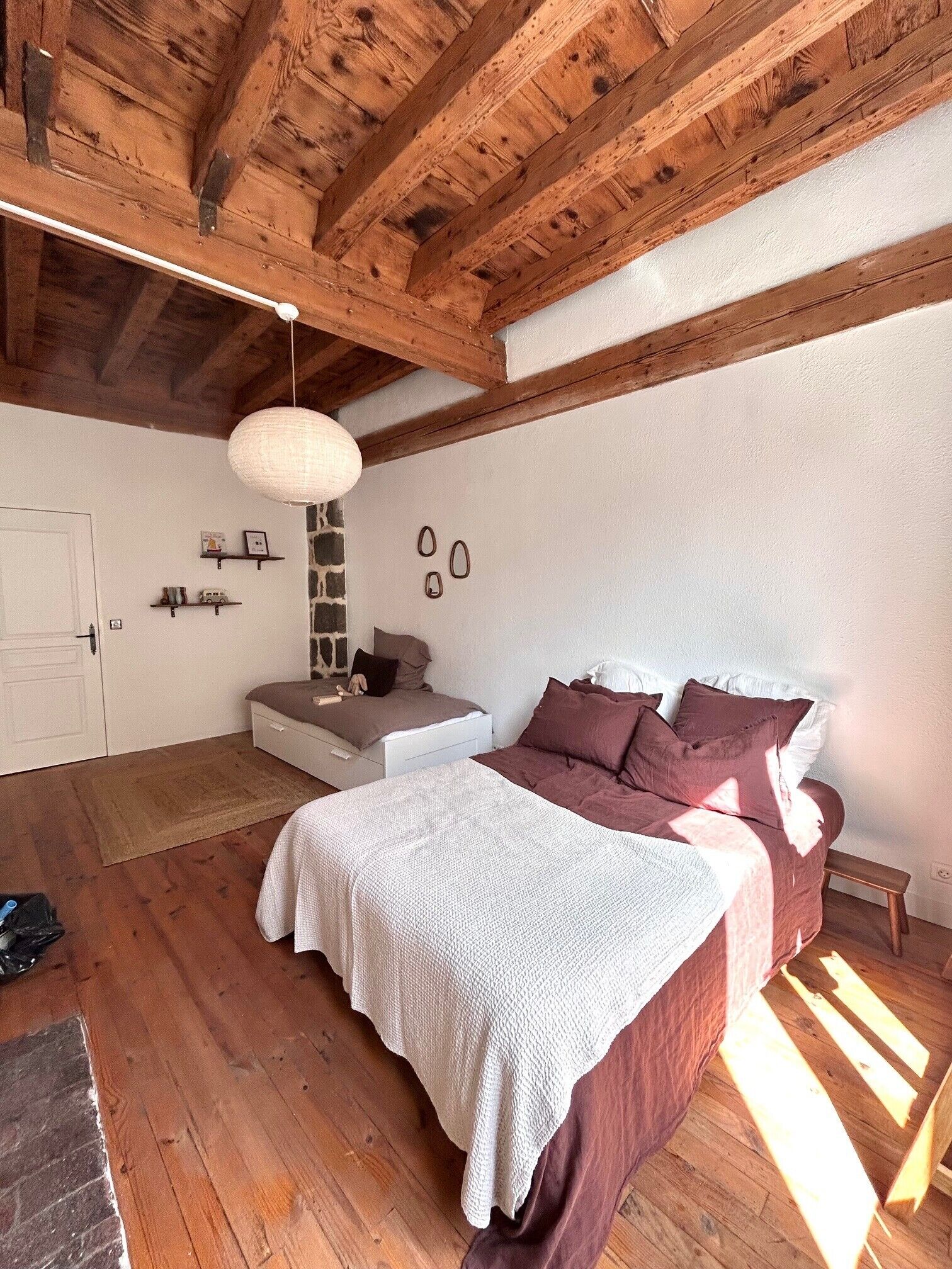 Quarto com teto de madeira, cama com colcha branca e castanha, mesa de cabeceira e luminária pendente.