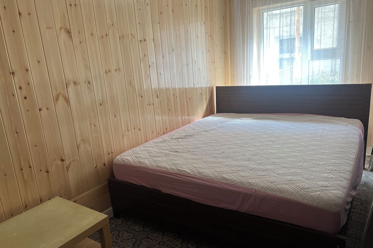 Ein Schlafzimmer mit holzverkleideter Wand, einem Bett mit gesteppter Matratze, einem Nachttisch und einem Fenster mit Vorhängen.