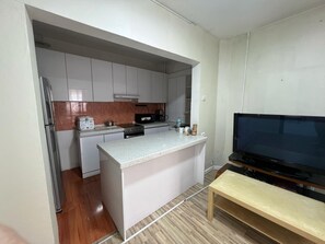 Cocina compacta con isla integrada, refrigerador, estufa y microondas. Hay un banco y un televisor en la sala.