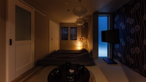 A dimly lit room with a bed, a small table with bowls and chopsticks, a wall-mounted TV, and a patterned wallpaper. - Oyado Kinokuniya Takaramachi (Nagasaki)