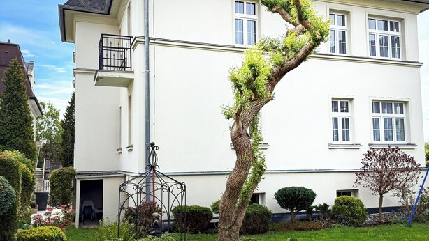 A two-story house with a white exterior and dark roof. There is a small balcony on the second floor and a garden with various plants and a tree.