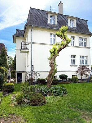 Una casa de dos pisos con fachada blanca y techo oscuro. Tiene un pequeño balcón en el segundo piso y un jardín con diversas plantas y un árbol.