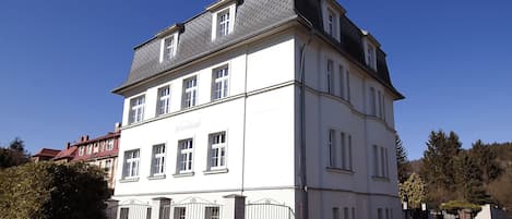 A large, white, multi-story building with a dark roof, several windows, and a gated entrance.
