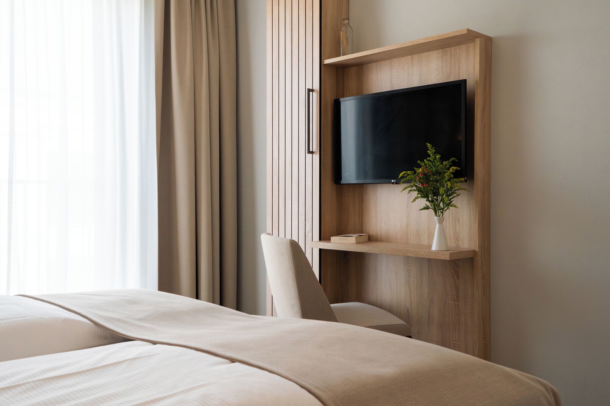 Quarto de hotel moderno com uma cama, uma TV montada e uma pequena estante com uma planta.