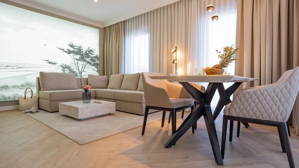 Una sala de estar moderna con un sofá grande, una mesa de comedor y sillas. Se ve la playa a través de amplios ventanales.