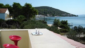 A terrace with a table and two red chairs, overlooking a coastal area with a hill and a body of water.