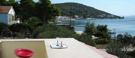A terrace with a table and two red chairs, overlooking a coastal area with a hill and a body of water.