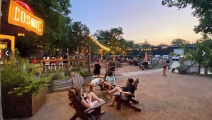 An outdoor seating area with wooden chairs and tables, surrounded by greenery and string lights.