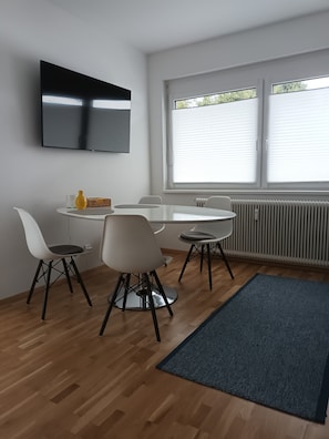 Un espace repas moderne avec une table ronde, des chaises blanches, un téléviseur à écran plat fixé au mur et une fenêtre avec des stores.