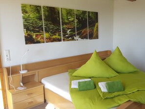 Un dormitorio con una cama, almohadas verdes, una mesita de noche, un cuadro en la pared y un enchufe.