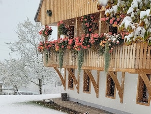 Um edifício de madeira com uma varanda decorada com flores, chão coberto de neve e uma árvore ao fundo.