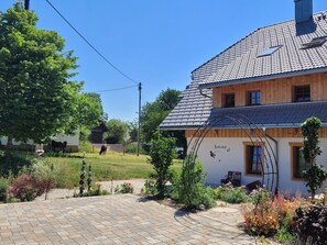 Uma casa de dois andares com telhado em telhas, um jardim com diversas plantas e um caminho de entrada pavimentado.